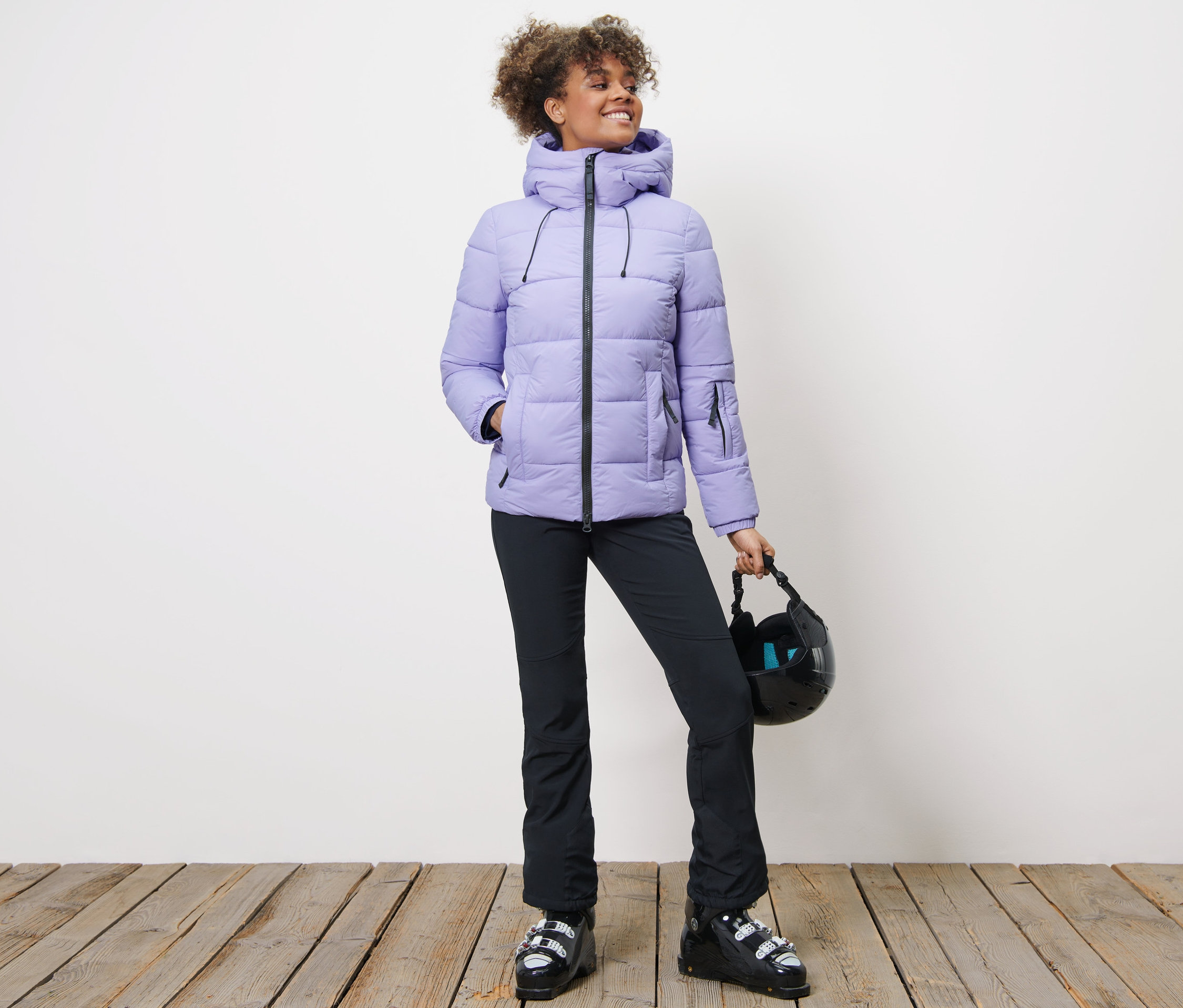 Frau in Ski-Steppjacke, schwarzer Hose und Skischuhen hält einen Helm.