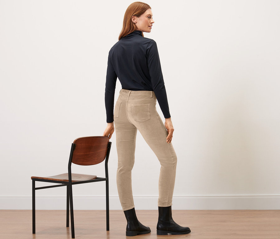 Frau steht seitlich und stützt sich auf einem Stuhl, sie trägt eine Samthose in Camel.