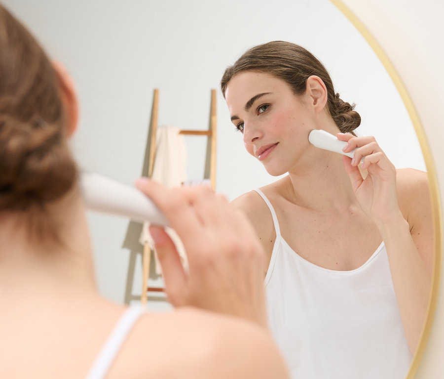 Frau benutzt einen elektrischen Gesichtshaarentferner vor einem Spiegel.