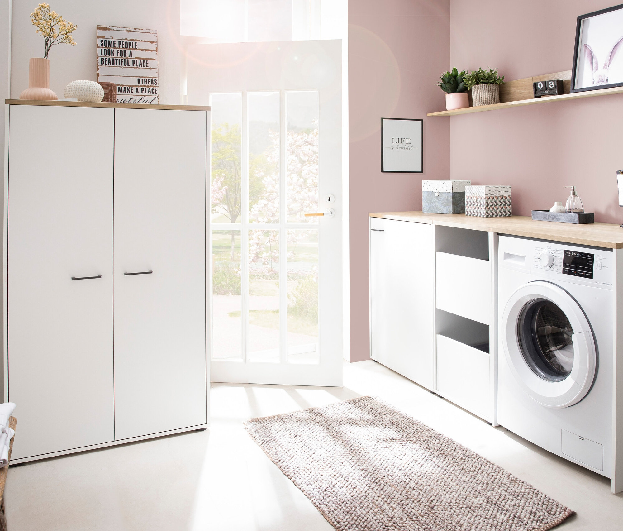 Einrichtung eines Waschraums mit einer weißen Waschmaschine, Schränken und einer offenen Tür. Zu sehen ist ein Mehrzweckschrank »Solea« in matt weiß.