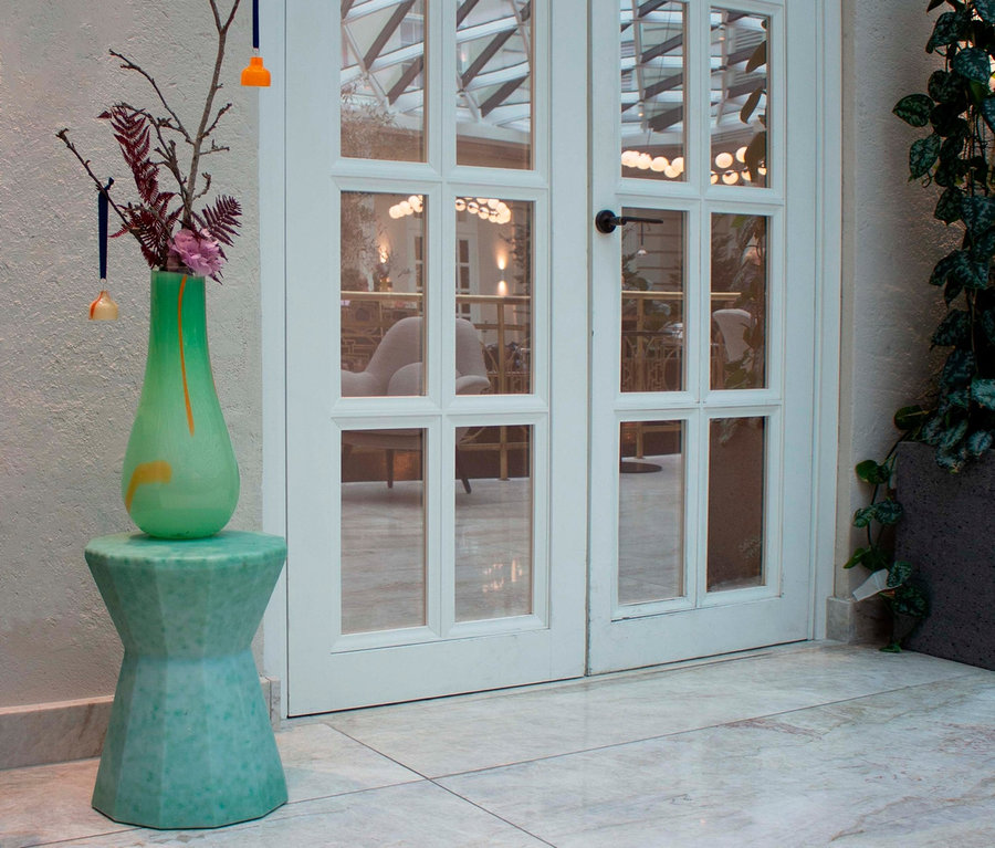 Blumen in Vase auf grünem Beistelltisch vor Glastür.