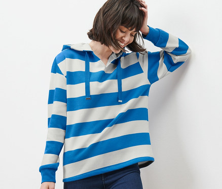 Frau lehnt an Wand und trägt einen Gestreiften Hoodie, Jeans und Turnschuhe.