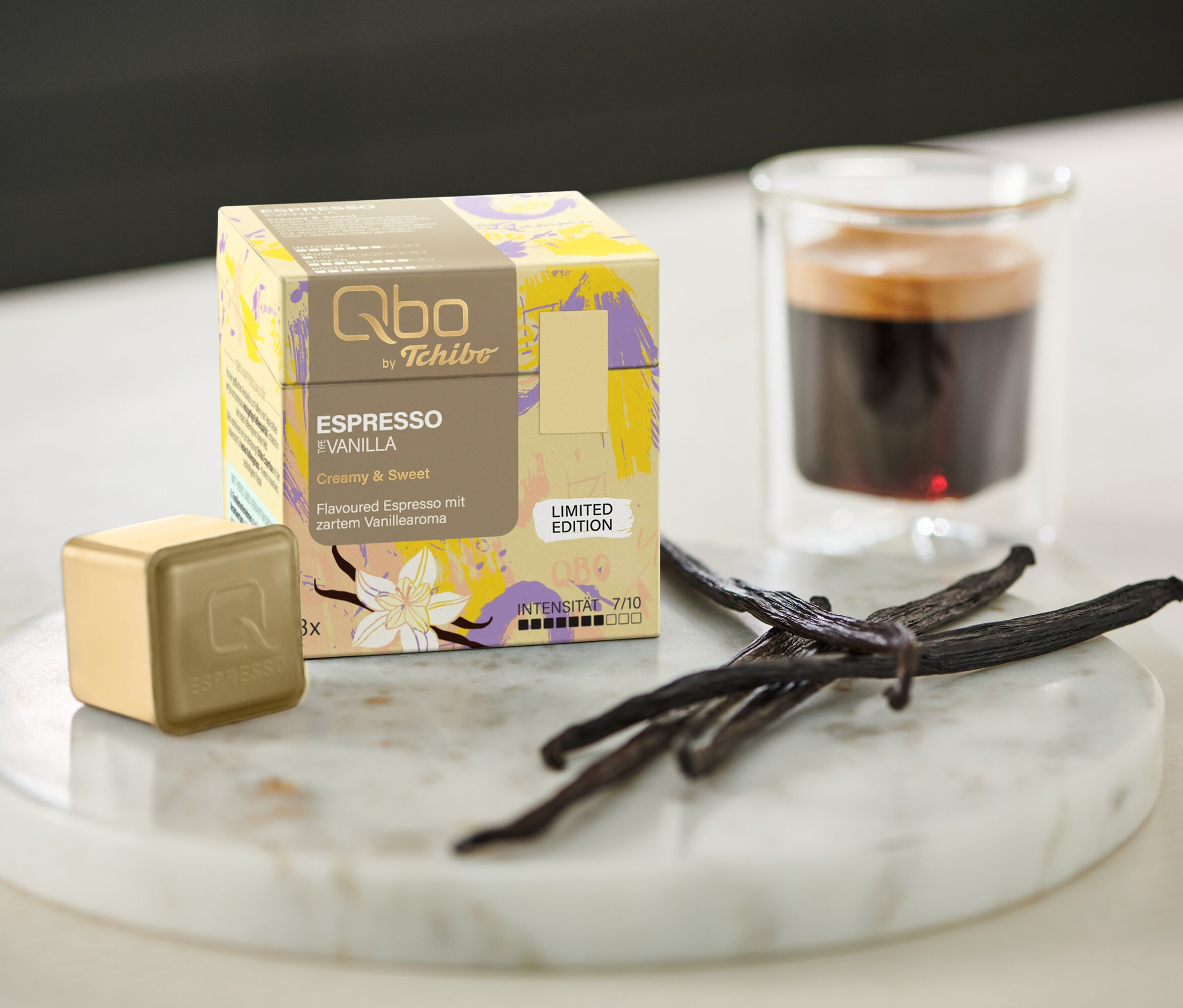 Eine Qbo Flavoured Espresso Vanilla - 8 Kapseln Packung, Kapsel, Espresso und Vanilleschoten auf einem Marmortablett neben einem Glas.