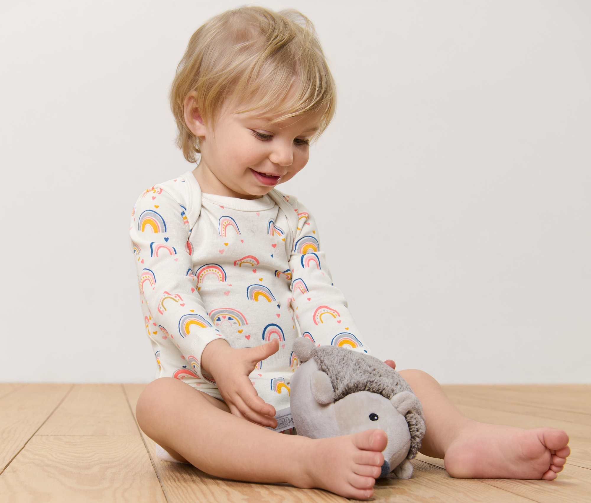 Ein Kind sitzt auf dem Boden und spielt mit einem Plüschigel. Es trägt einen Langarmbody mit Regenbogenmuster.