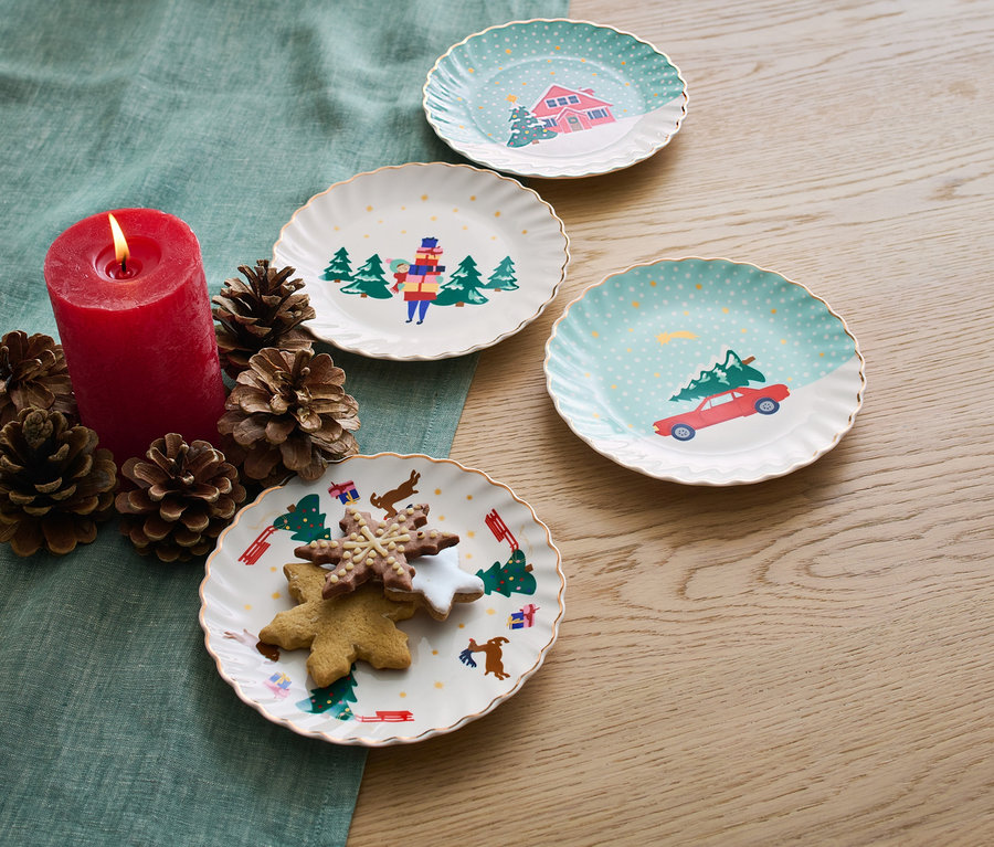 Vier Dessertteller mit Glanzgolddekor, Kerze, Zapfen und Kekse auf einem Tisch.