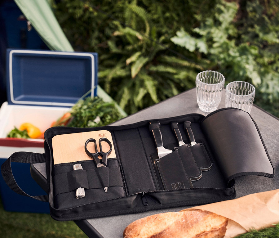 Offenes schwarzes ZWILLING Messer-Traveller-Set mit Holzbrett und Schere auf einem Tisch neben einer Kühlbox.
