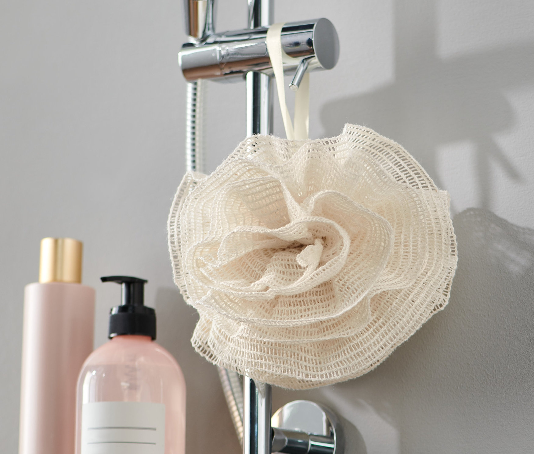 Ein Duschschwamm aus Naturfaser hängt an einer Duschstange neben zwei Flaschen.