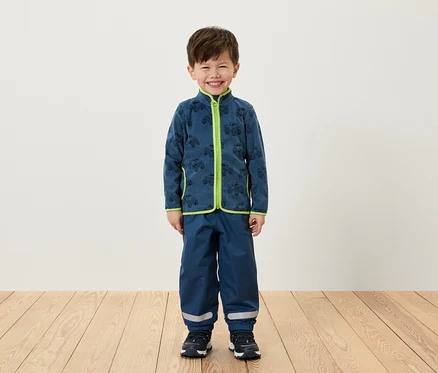 Ein Junge steht und trägt eine blaue Fleecejacke mit grünen Akzenten und eine blaue Hose.