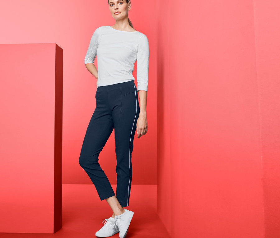 Frau posiert im Studio vor rotem Hintergrund und trägt eine Stretchhose, ein weißes T-Shirt und Turnschuhe.