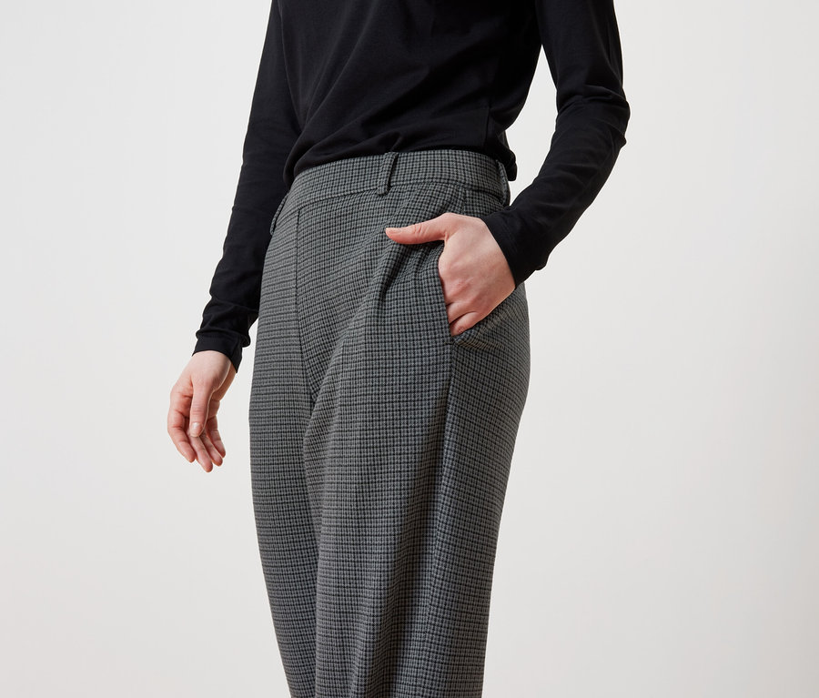 Eine Frau hat ihre Hand in der Tasche einer grauen, karierten Hose und trägt ein schwarzes Langarmshirt.