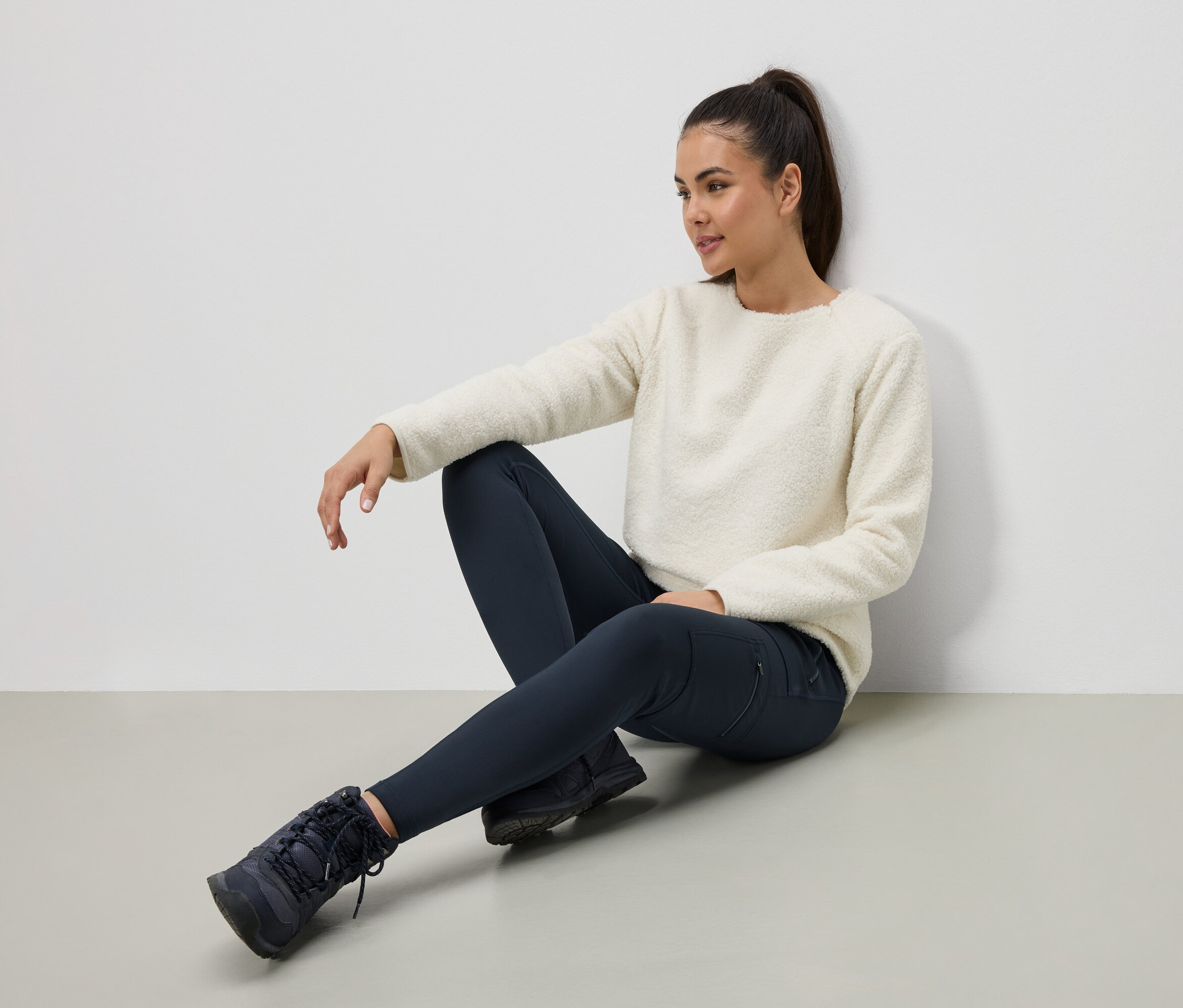 Frau sitzt auf dem Boden und trägt einen cremefarbenen Pullover, dunkelblaue Leggings und Stiefel.