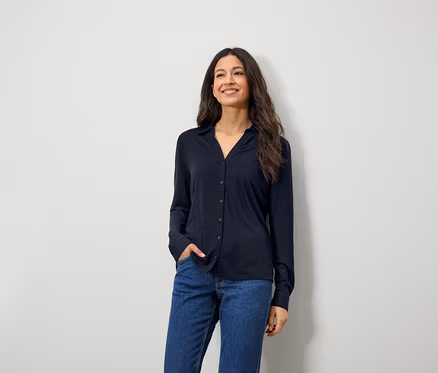 Eine lächelnde Frau mit langen braunen Haaren posiert in einem blauen Langarmhemd und Jeans.