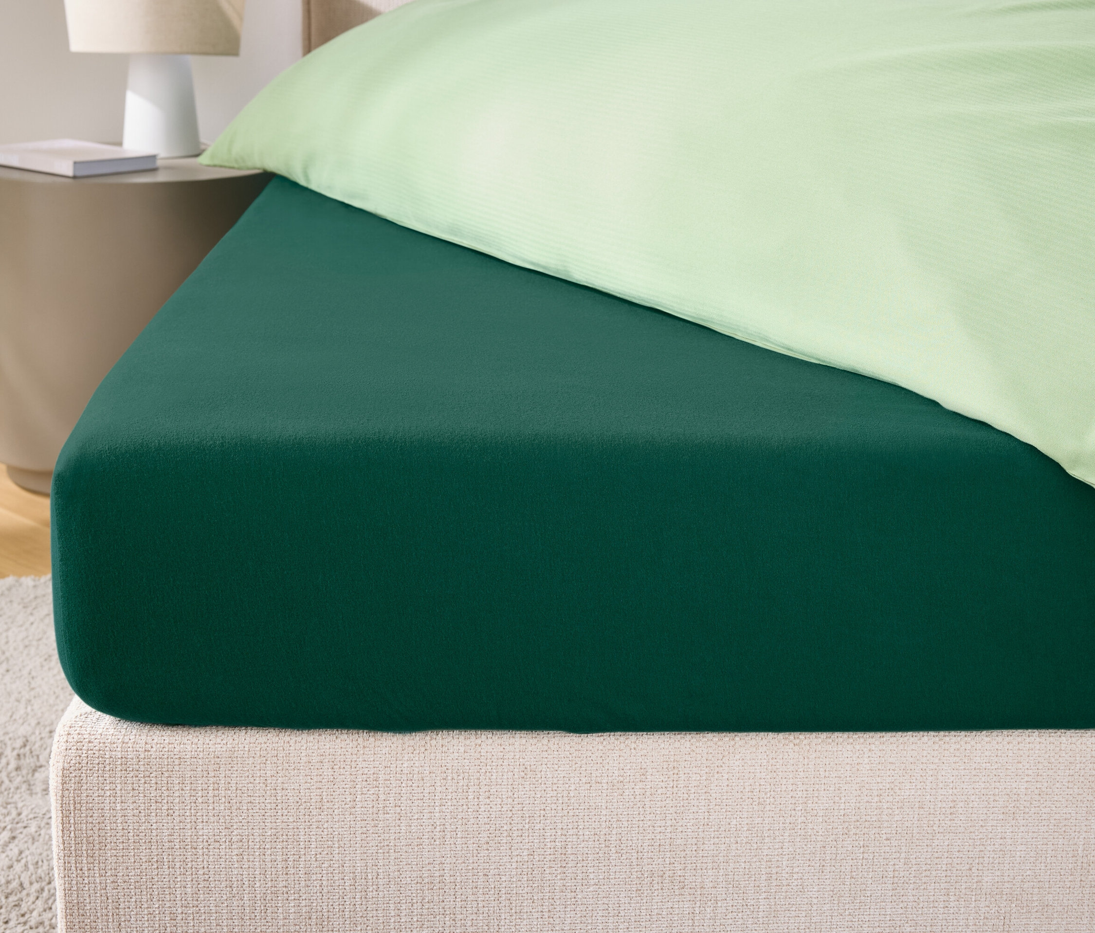 Detailaufnahme eines grünen Spannbettlakens auf einem Bett mit cremefarbenem Kopfteil und hellgrünem Kissenbezug.