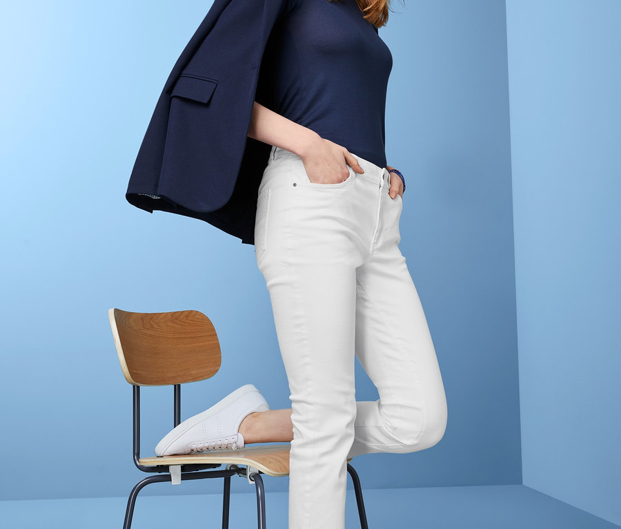 Eine Frau posiert in weißen Straightfit-Jeans, einem marineblauen Oberteil und einem Blazer über der Schulter, während sie sich an einen Stuhl lehnt.