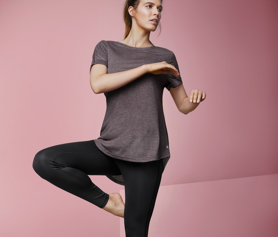 Frau in Sportkleidung steht auf einem Bein und trägt ein Longshirt.