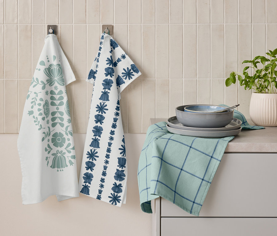 Zwei Geschirrtücher mit floralen Mustern hängen nebeneinander an der Wand. Darunter liegt ein gefaltetes grünes Geschirrtuch mit blauen Streifen, auf dem Teller und eine Schüssel gestapelt sind. Daneben steht ein Topf mit Kräutern.