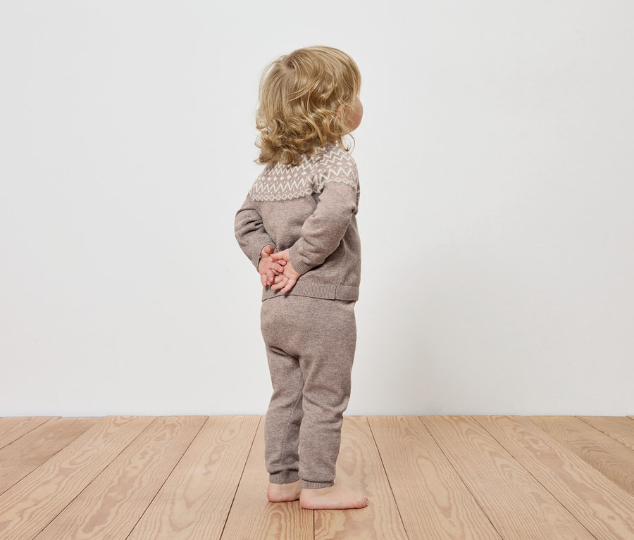 Ein Kind in brauner Strickkleidung steht mit dem Rücken zur Kamera auf einem Holzboden.