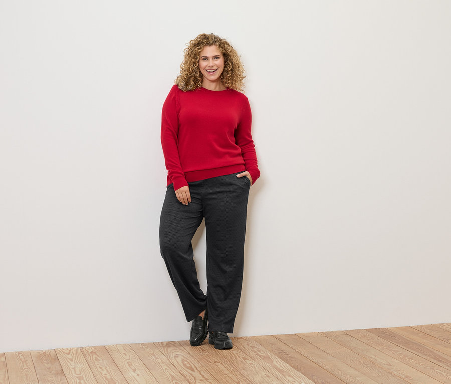 Eine Frau mit lockigem Haar lehnt an einer Wand und trägt einen roten Pullover und eine dunkle Hose.