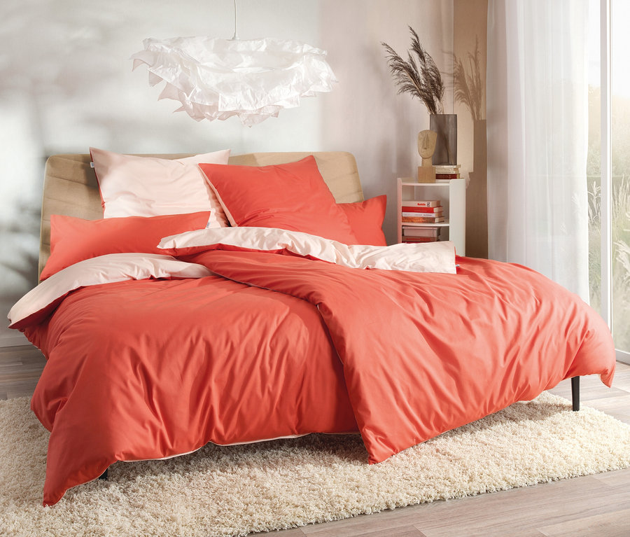 Bett mit korallenroter SCHÖNER WOHNEN-Kollektion Bettwäsche, Übergröße und Normalgröße auf einem weißen, zotteligen Teppich.