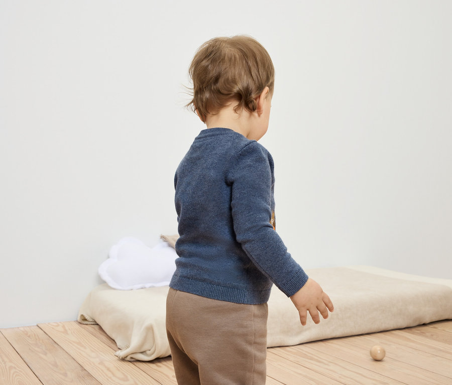 Kleinkind in blauem Pullover und brauner Hose steht mit dem Rücken zur Kamera und spielt mit einem Holzspielzeug.