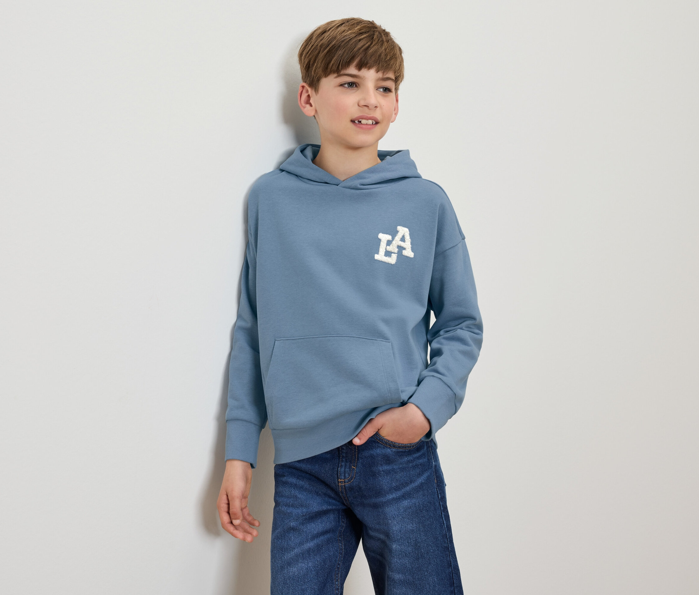 Ein Junge steht und posiert in einem blauen Kapuzenpullover mit einem weißen LA-Logo und dunkelblauen Jeans.