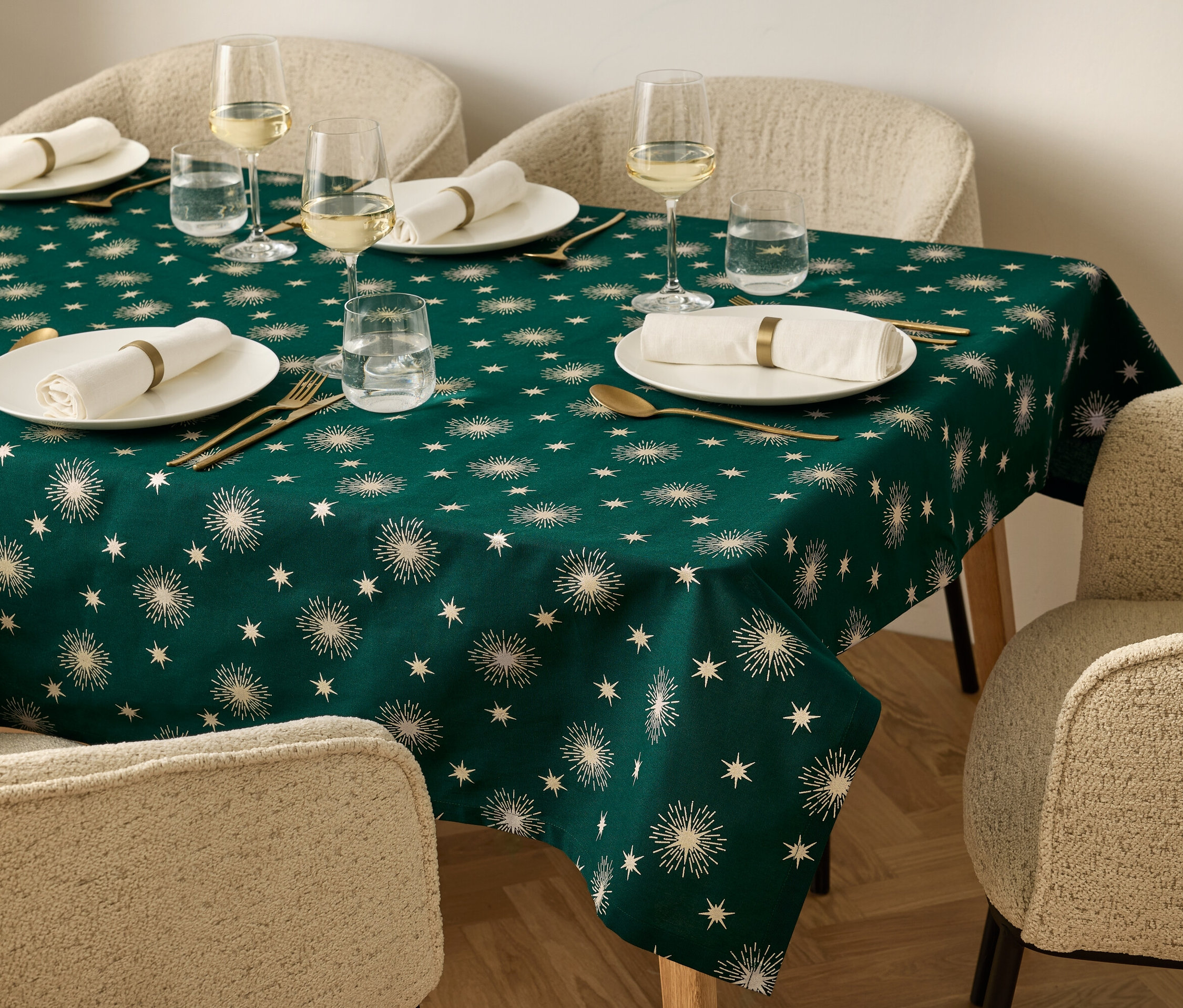 Ein gedeckter Tisch mit einer dunkelgrünen Tischdecke mit Sternenmuster, Tellern, Gläsern, goldenem Besteck und beigen Stühlen.