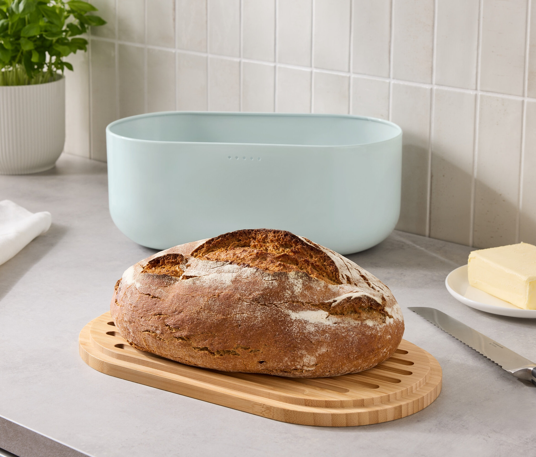 Ein Brotlaib auf einem Holzbrett neben einem blauen Brotkasten, einem Stück Butter und einem Messer auf einer Küchentheke.