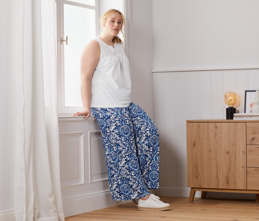 Frau lehnt am Fenster in Top mit Stickerei und bedruckter Palazzohose.