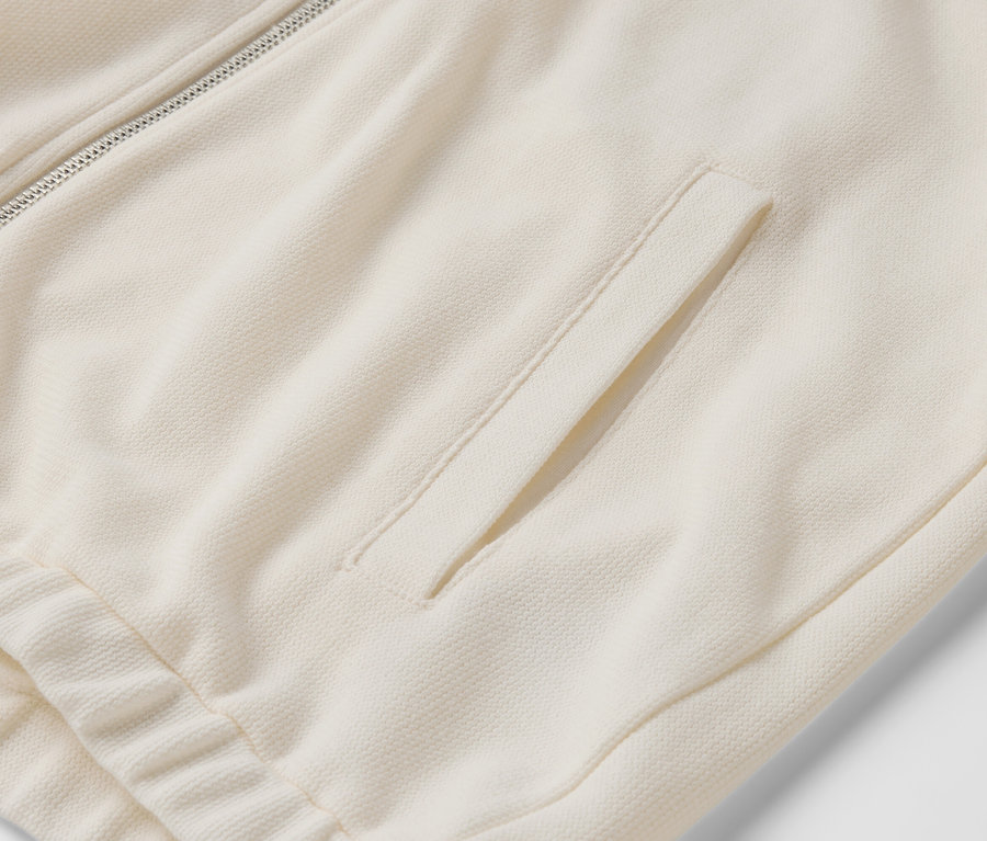 Detailansicht einer cremefarbenen Trainingsjacke mit Reißverschluss und Tasche.