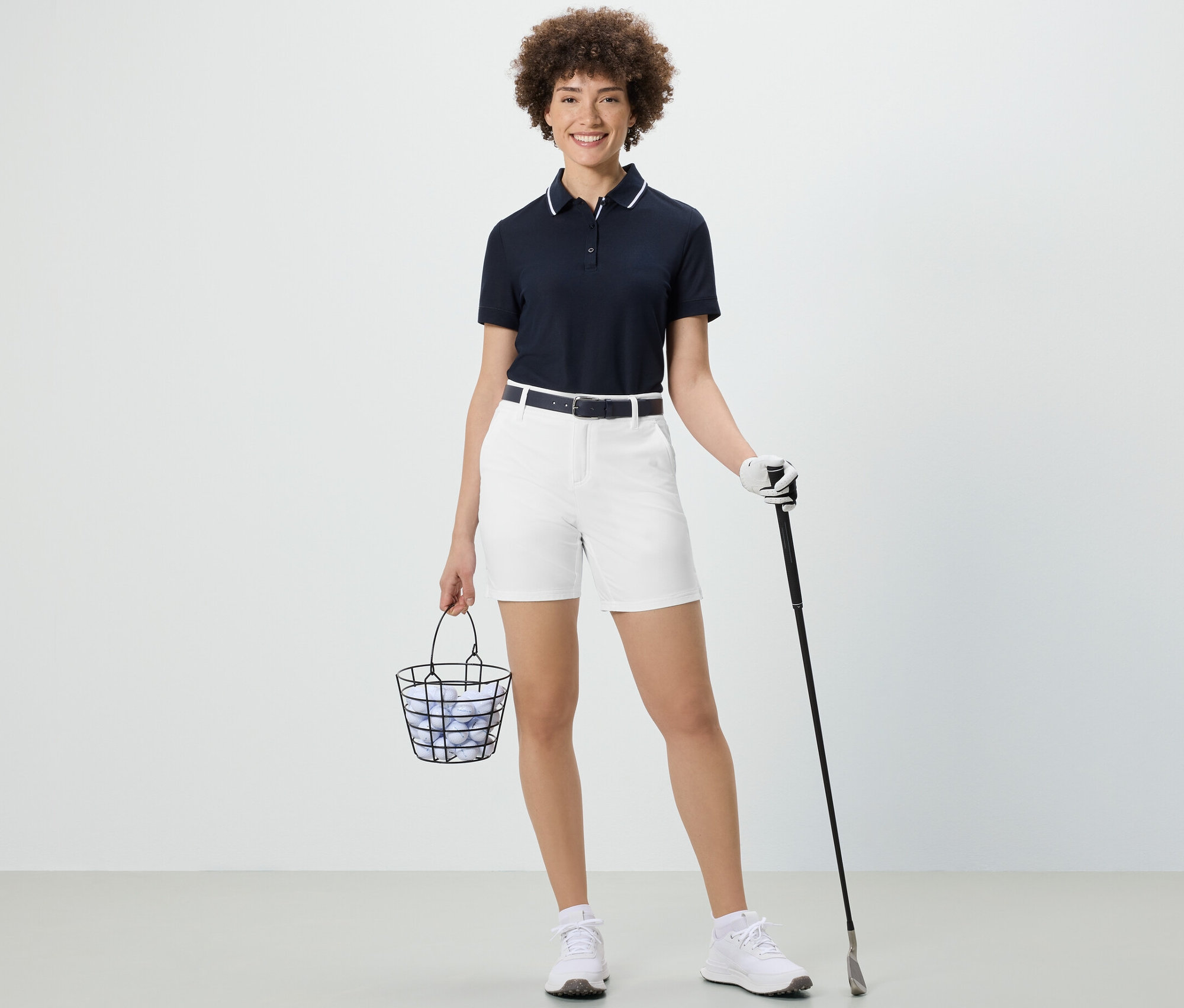 Frau hält einen Korb mit Golfbällen und einen Golfschläger. Sie trägt weiße Softshell-Funktionsshorts.