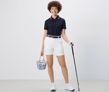 Frau hält einen Korb mit Golfbällen und einen Golfschläger. Sie trägt weiße Softshell-Funktionsshorts.