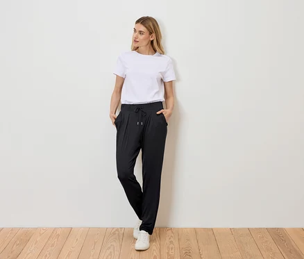 Frau steht mit Händen in den Taschen vor einer Wand, trägt ein weißes T-Shirt, eine schwarze Hose mit Kordelzug und weiße Turnschuhe.