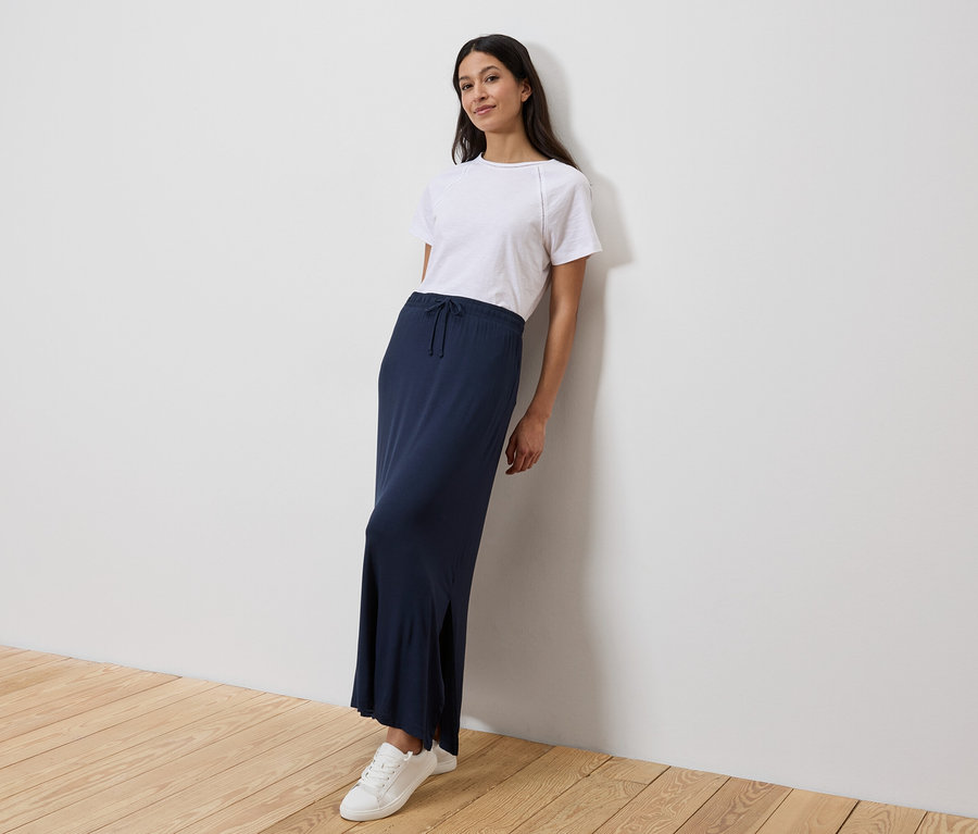 Frau lehnt an einer Wand, trägt ein weißes T-Shirt, einen langen, marineblauen Rock und weiße Turnschuhe.