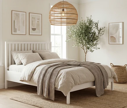 Schlafzimmer mit weißem Bett, beiger Bettwäsche und Überwurf. Über dem Bett hängen zwei Bilder, daneben steht ein Olivenbaum.