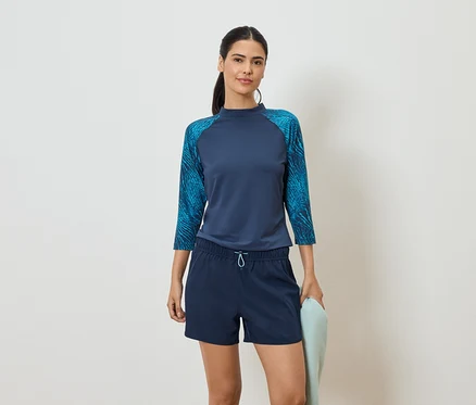 Frau mit marineblauem Oberteil mit Dreiviertelärmeln und blauen Shorts, hält ein hellblaues Handtuch vor einem weißen Hintergrund.