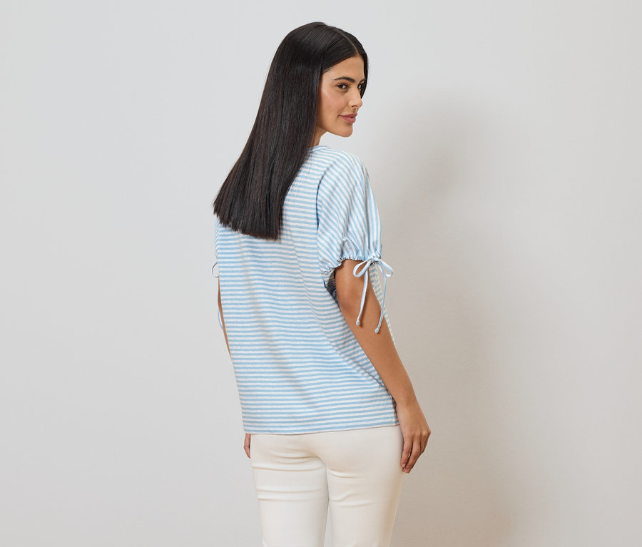 Eine Frau mit langen, dunklen Haaren trägt ein hellblau-weiß gestreiftes T-Shirt mit Bindebändern an den Ärmeln und eine helle Hose.