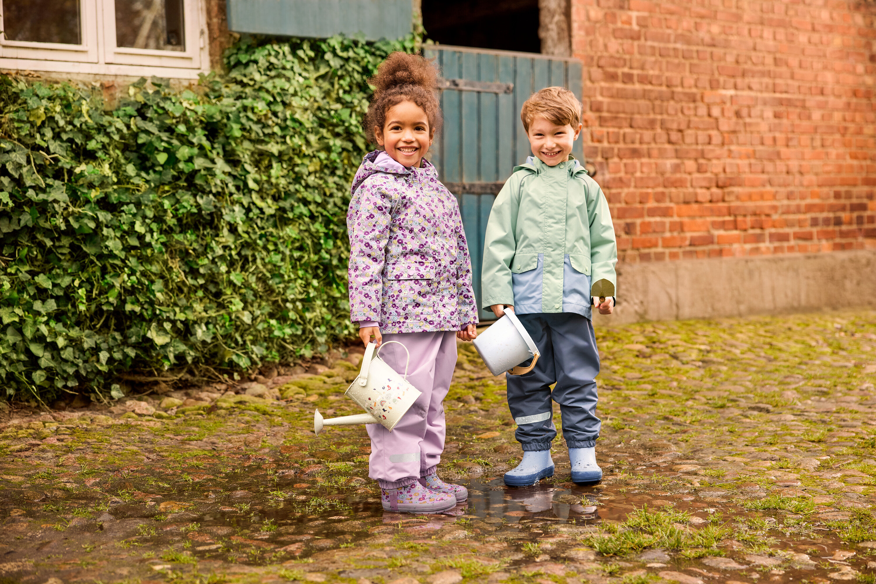 Zwei Kinder stehen draußen: ein Mädchen mit einer geblümten Kinder-Regenjacke und geblümten Kinder-Regenstiefeln, und ein Junge mit einer grünen Kinder-Regenjacke und blauen Kinder-Regenstiefeln. Beide tragen Kinder-Regenhosen.