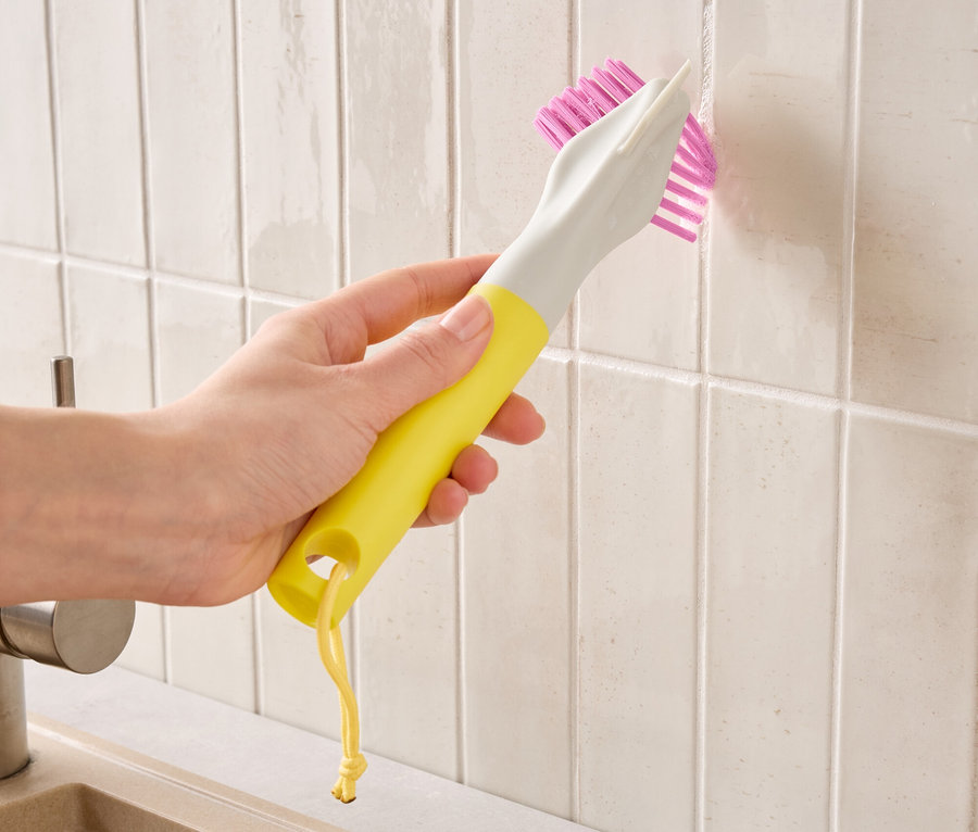Eine Hand hält eine gelb-graue Bürste mit pinken Borsten und reinigt Fliesenfugen.
