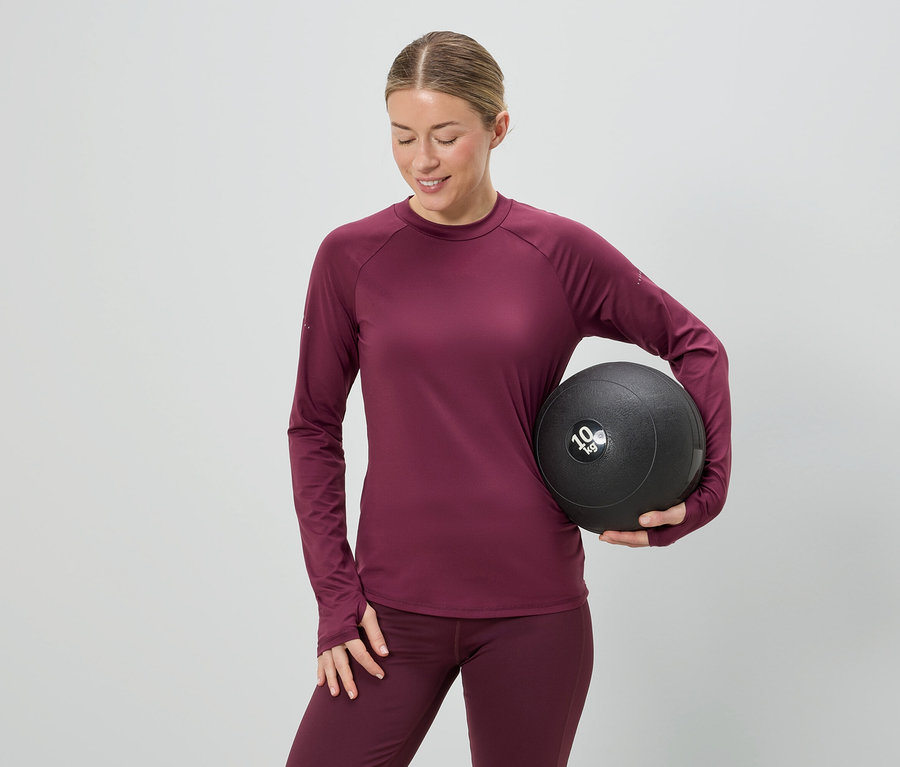 Frau in bordeauxfarbenem Sportoutfit hält einen 10 kg schweren Medizinball vor einem grauen Hintergrund.