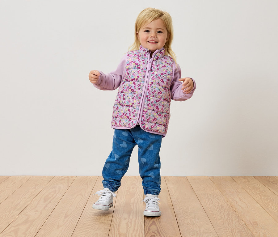 Ein kleines blondes Mädchen steht auf einem Holzboden und trägt eine Blumenweste, ein lila Oberteil und Jeans.