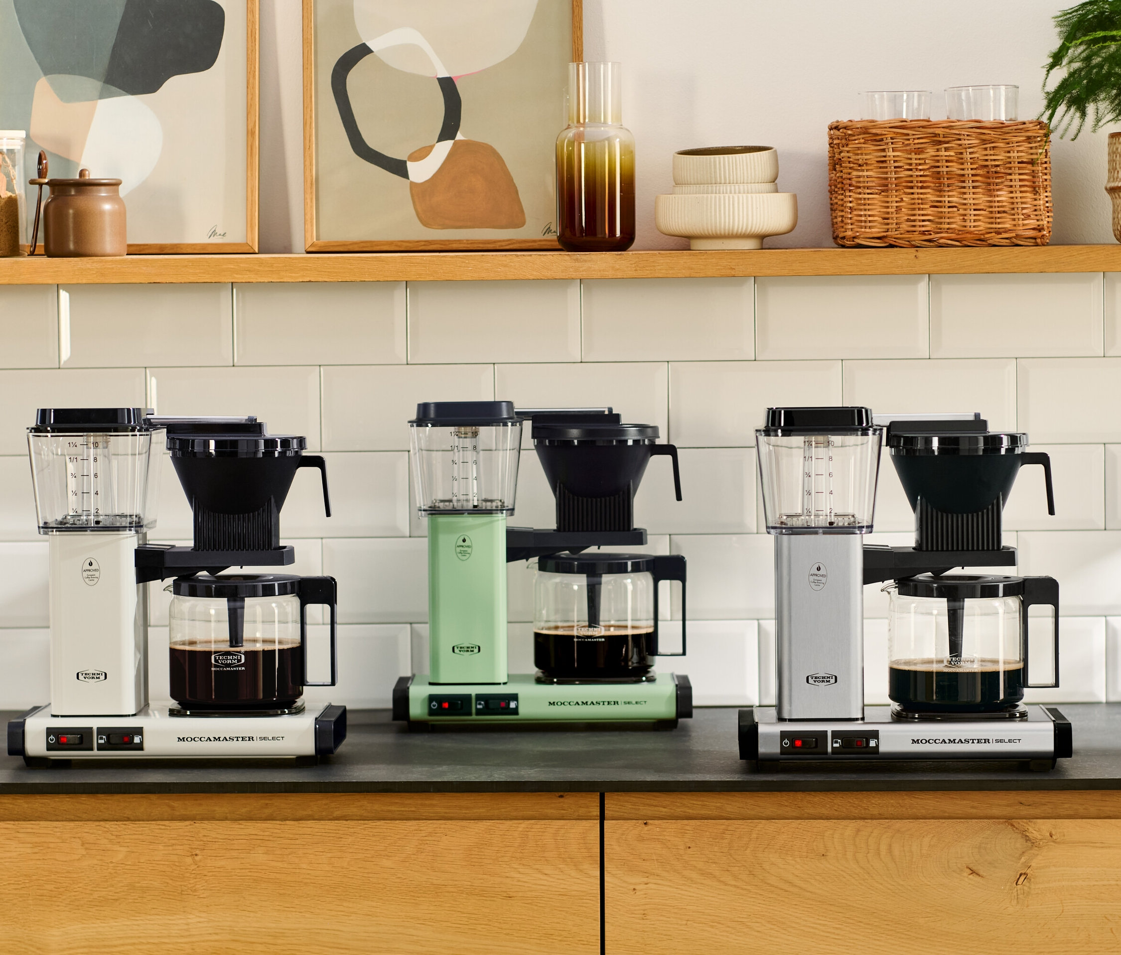 Drei Moccamaster KBG Select Filterkaffeemaschinen in Weiß, Grün und gebürstetem Alu stehen auf einer Küchentheke.