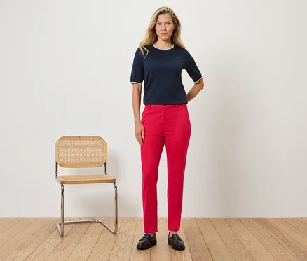 Eine Frau steht vor einer Wand neben einem Stuhl. Sie trägt eine marineblaue Bluse mit weißen Details und eine rote Hose.