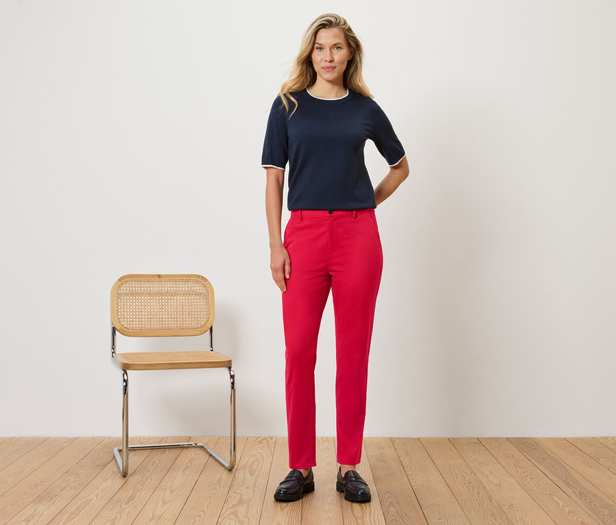 Eine Frau steht vor einer Wand neben einem Stuhl. Sie trägt eine marineblaue Bluse mit weißen Details und eine rote Hose.