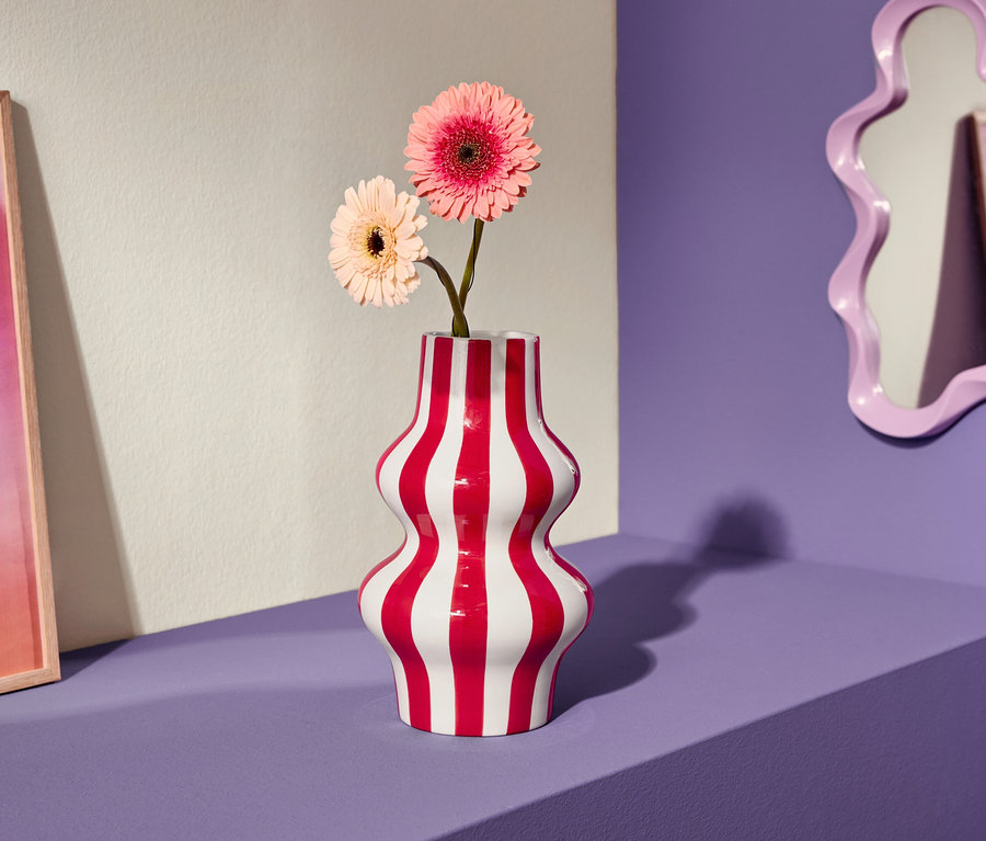 Zwei rosa Gerbera-Blumen in rot-weiß gestreifter Vase auf lila Regal, daneben ein pinkfarbenes Bild und ein geschwungener Spiegel.