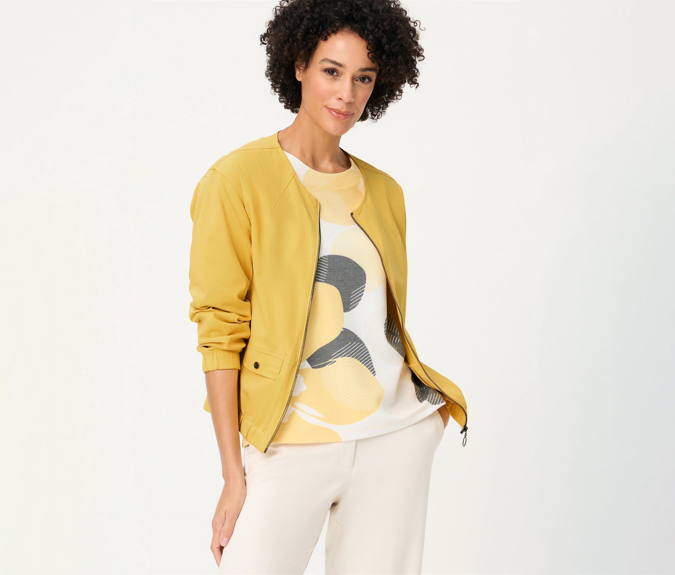 Frau mit lockigem Haar trägt eine gelbe Olsen Sweatjacke über einem gemusterten Oberteil und einer hellen Hose.