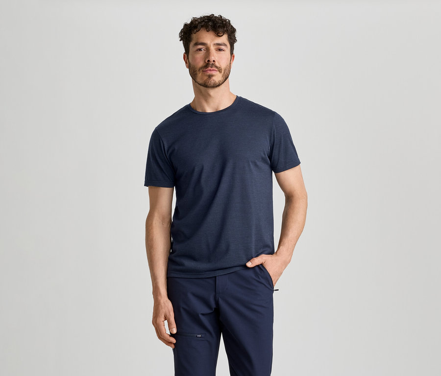 Ein Mann mit lockigem Haar und Bart trägt ein dunkelblaues T-Shirt und eine dunkelblaue Hose. Er steht mit der rechten Hand in der Hosentasche.