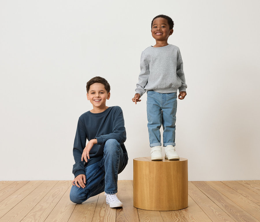 Zwei Jungen posieren: Einer hockt in einem marineblauen Pullover und Jeans, der andere steht in einem grauen Pullover und Jeans auf einem Holzhocker.
