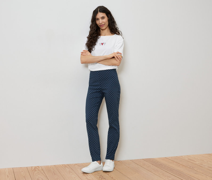 Frau steht mit verschränkten Armen, trägt ein weißes T-Shirt mit Herzen und blaue Leggings mit weißen Punkten.