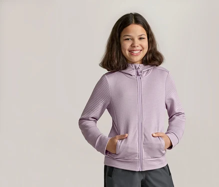 Lächelndes Mädchen mit den Händen in den Taschen einer lila Kapuzenjacke mit Reißverschluss und grauer Hose.