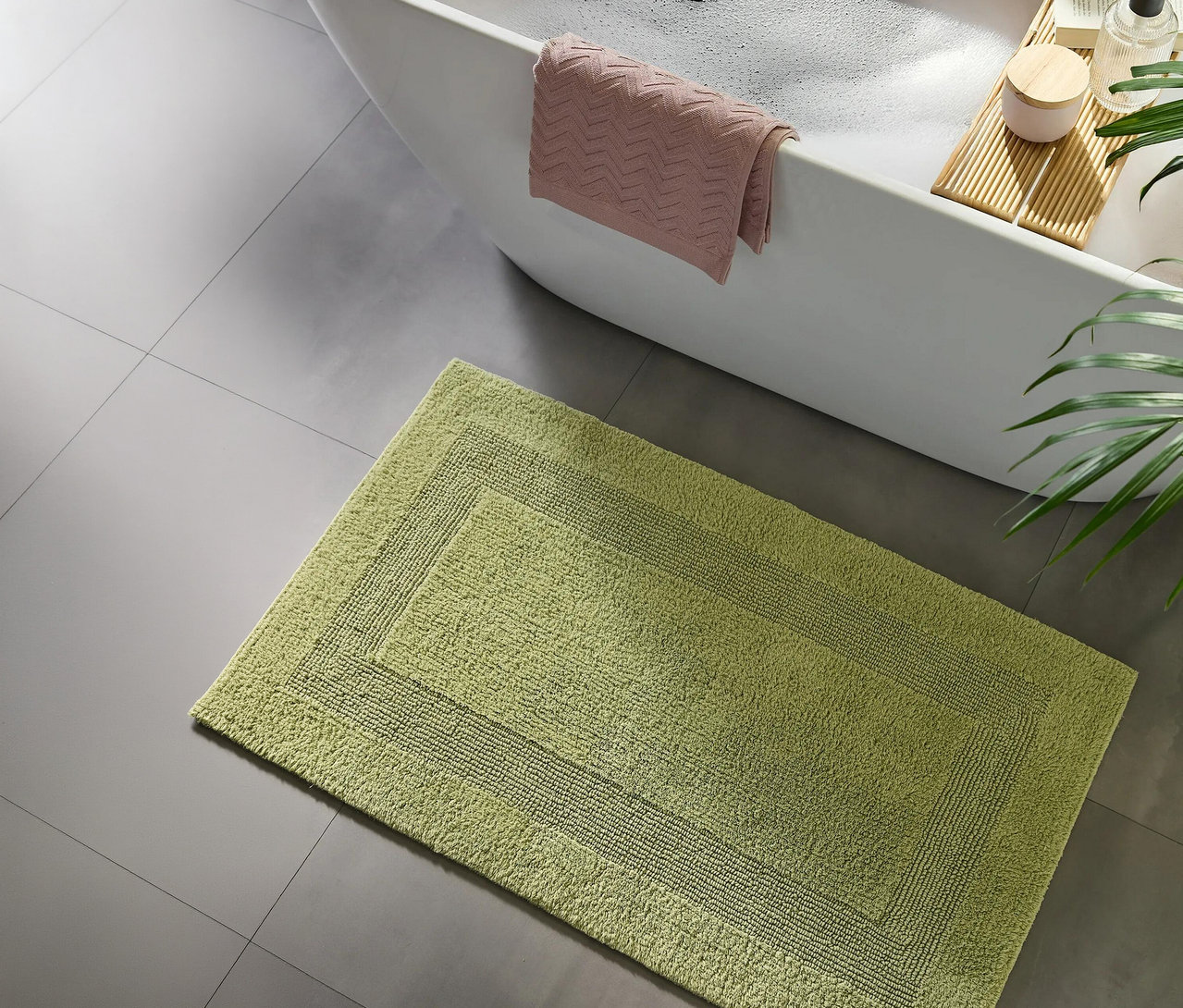 Ein grüner Badteppich liegt auf grauem Fliesenboden neben einer weißen Badewanne, über die ein rosa Handtuch hängt. Ein Holzbadebrett mit Kerze und Flasche liegt auf der Wanne, rechts ist eine Pflanze zu sehen.